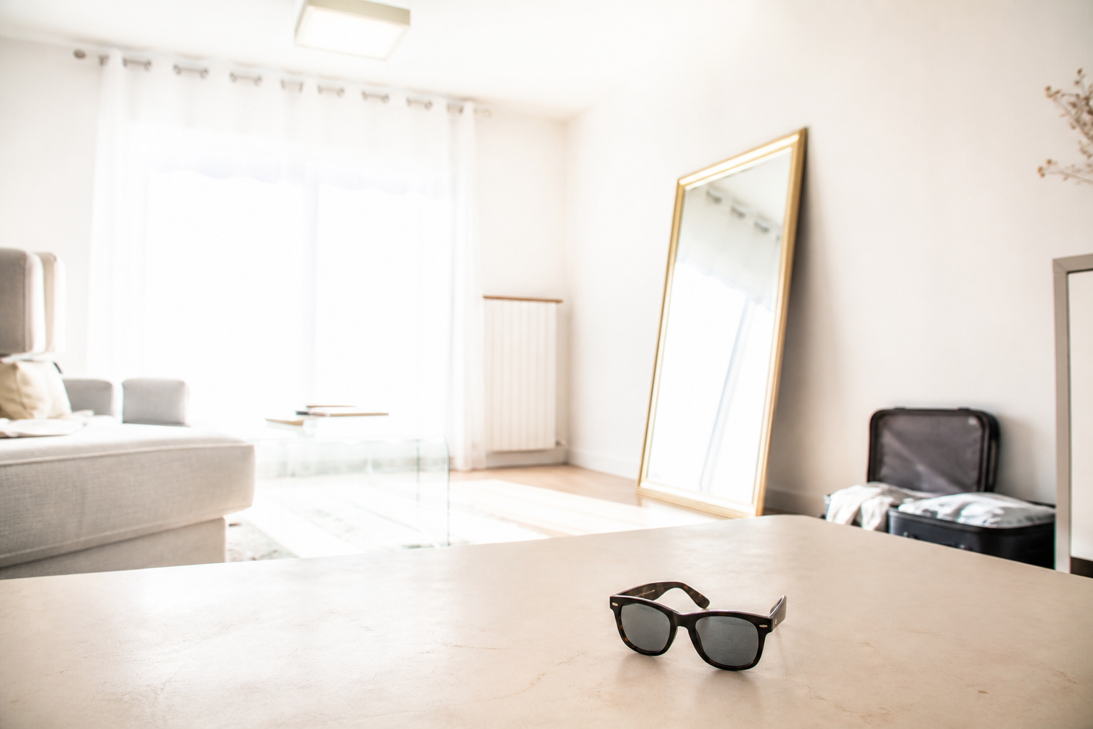Salon trop blanc, voilages, miroir, LED, lunettes noires et valise ouverte.